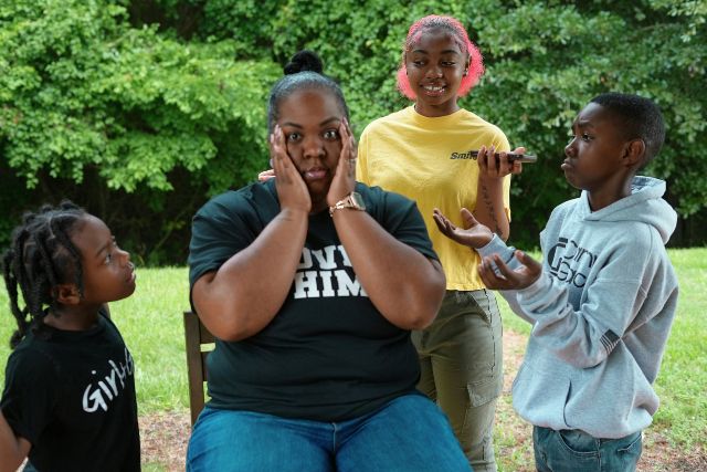 A woman seated outdoors reacts with surprise as three children playfully engage with her in a grassy setting | Pathway Caring for Children