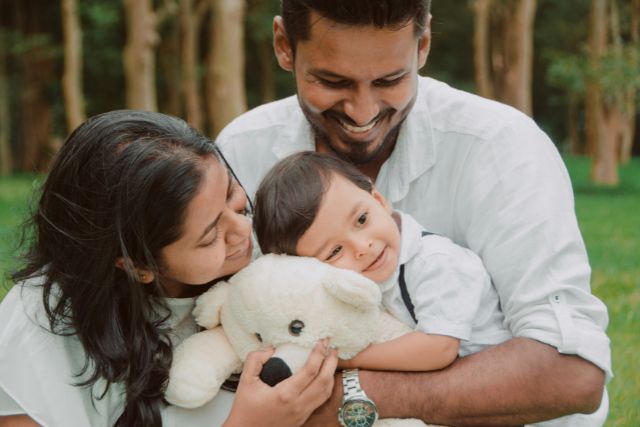 A couple sits outdoors in the grass smiling and embracing their young child who holds a stuffed animal | Pathway Caring for Children