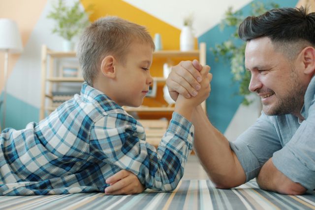 A man and a young boy smile while arm wrestling on the floor of a colorful living room | Pathway Caring for Children