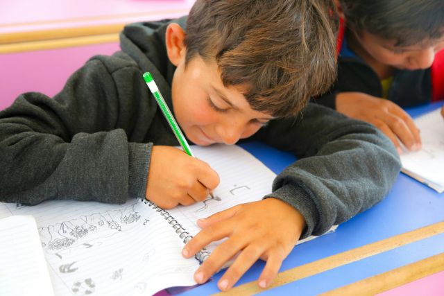 A boy in a dark jacket writes with a green pencil on a worksheet at a desk | Pathway Caring for Children
