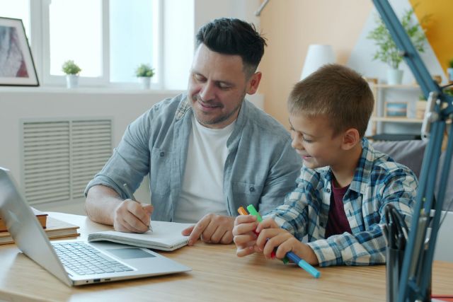 A man and a young boy sit side by side at a table, looking at a laptop and papers together | Pathway Caring for Children