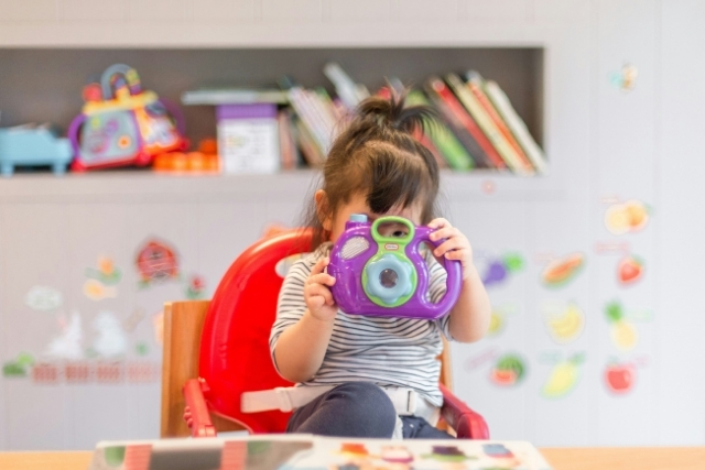A toddler in a high chair plays with a toy camera surrounded by books and toys | Pathway Caring for Children