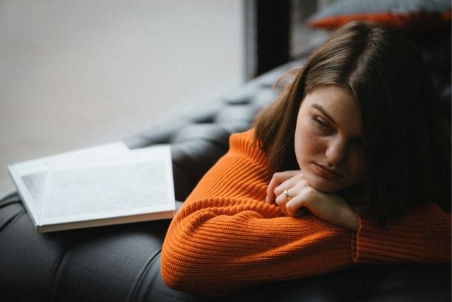 A girl in an orange sweatshirt rests her head on her arm beside an open laptop on a dark couch | Pathway Caring for Children