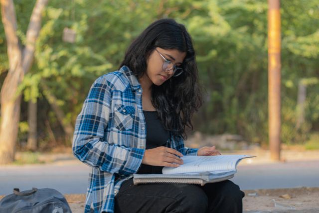 A teenage girl in a blue plaid shirt sits outdoors with a notebook in her lap, surrounded by trees | Pathway Caring for Children