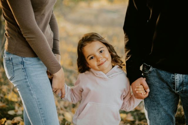 A young girl smiles at the camera while holding the hands of two adults walking together along an autumn path | Pathway Caring for Children